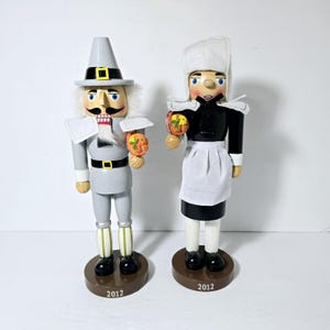 May include: A pair of wooden pilgrim nutcrackers, one in gray and one in black, both holding a basket of gourds. The bases of the nutcrackers are brown and have the year 2012 printed on them.