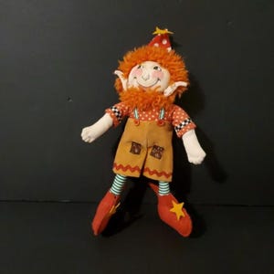 Könnte beinhalten: Eine Plüschpuppe aus orangefarbenem und braunem Stoff, die einen Elfen mit einem rot-weißen Hut und einem Stern obenauf darstellt. Die Puppe trägt ein rot-weiß gepunktetes Hemd und eine braune Hose mit einem grün-weiß gestreiften Saum. Die Puppe hat einen rot-weiß gestreiften Saum an den Schuhen.
