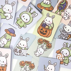 May include: A collection of colorful, illustrated cards featuring adorable white bunny characters in various costumes and settings. The cards showcase themes like Halloween, space, and aliens, with soft pastel backgrounds and cute designs.