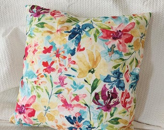 May include: Square throw pillow with a vibrant floral pattern. The design features watercolor-style flowers in shades of red, pink, yellow, blue, and purple, set against a white background. Green leaves and stems complete the botanical theme.