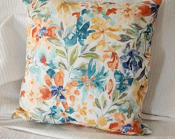 May include: Square throw pillow with a floral pattern. The pillow features a white background with a watercolor design of orange, blue, teal, and yellow flowers and green leaves. The pillow is likely made of a soft fabric.