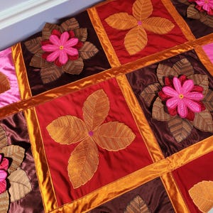 May include: A patchwork quilt with a floral design. The quilt is made of different colored fabrics, including red, orange, brown, and pink. The flowers are embroidered with intricate details.