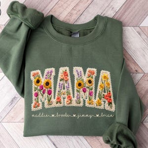 May include: Military green sweatshirt with the word "MAMA" embroidered with colorful flowers. The names "maddie", "brooker", "jimmy", and "brian" are embroidered below the word "MAMA". The sweatshirt has a crew neck and long sleeves.