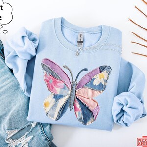 May include: A light blue crewneck sweatshirt featuring a large butterfly graphic made of various fabric patches in shades of pink, purple, blue, and white with floral patterns. The sweatshirt is paired with distressed light blue denim jeans.