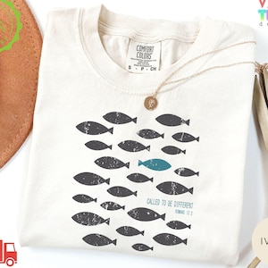 May include: Ivory-colored t-shirt with a crew neck, featuring a school of distressed gray fish, one teal fish, and the text "CALLED TO BE DIFFERENT" with a Bible verse reference. The shirt has a Comfort Colors tag.