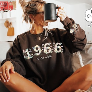 May include: A dark brown long-sleeve sweatshirt featuring the floral-patterned numbers 1966 on the front, with the text limited edition below. The left sleeve displays a small floral heart and the text chapter 9. A black mug is held in one hand.