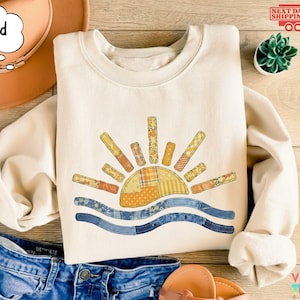 May include: Cream-colored sweatshirt featuring a colorful sun design with patchwork details. The sun has orange, yellow, and patterned rays, with blue waves below. The word "Sand" is in a speech bubble. The sweatshirt is paired with denim shorts.