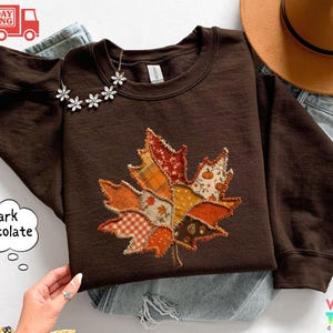 May include: A dark chocolate brown crewneck sweatshirt featuring a large, central maple leaf applique. The leaf is constructed from various fabric patches in orange, yellow, and white, displaying patterns of plaid, gingham, and small pumpkins.