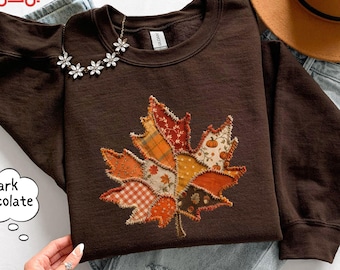 May include: A dark chocolate brown crewneck sweatshirt featuring a large, central maple leaf applique. The leaf is constructed from various fabric patches in orange, yellow, and white, displaying patterns of plaid, gingham, and small pumpkins.