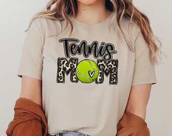 May include: A beige t-shirt with the text "Tennis Mom" in black lettering. The text is surrounded by a leopard print design and a green tennis ball with a white heart.
