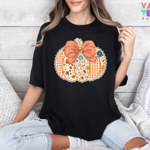 Może przedstawiać: Czarny t-shirt z wzorem dyni z kratkowaną kokardą i kwiatowymi akcentami. Dynia jest pomarańczowa z białym koronkowym wykończeniem. Tekst "VALENTEEN DESIGN" jest widoczny w prawym g&oacute;rnym rogu.