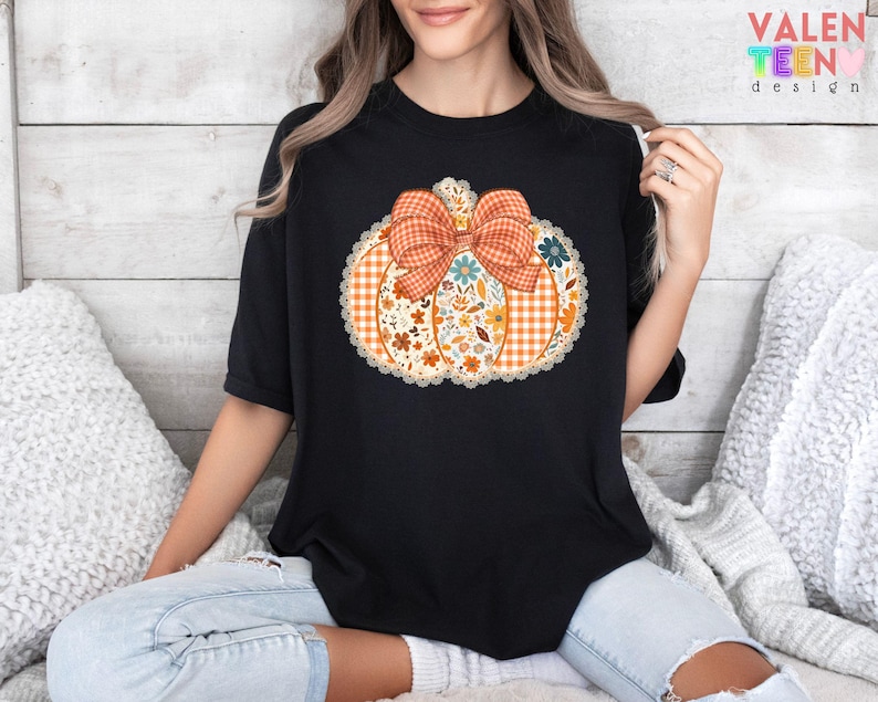 Może przedstawiać: Czarny t-shirt z wzorem dyni z kratkowaną kokardą i kwiatowymi akcentami. Dynia jest pomarańczowa z białym koronkowym wykończeniem. Tekst "VALENTEEN DESIGN" jest widoczny w prawym g&oacute;rnym rogu.