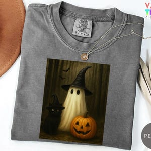 Puede incluir: Una camiseta gris Comfort Colors con un dise&ntilde;o de Halloween. El dise&ntilde;o muestra un fantasma con un sombrero de bruja, un gato negro y una calabaza tallada. Un collar plateado con un colgante est&aacute; sobre la camiseta.