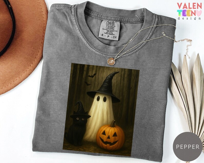 Puede incluir: Una camiseta gris Comfort Colors con un dise&ntilde;o de Halloween. El dise&ntilde;o muestra un fantasma con un sombrero de bruja, un gato negro y una calabaza tallada. Un collar plateado con un colgante est&aacute; sobre la camiseta.