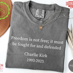 May include: A gray Comfort Colors t-shirt with the text "Freedom is not free; it must be fought for and defended" and "Charlie Kirk 1993-2025" in white. A gold necklace is draped on the shirt. The shirt has a round neckline.