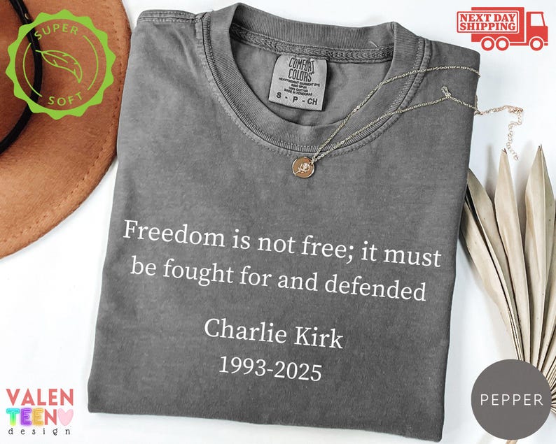 May include: A gray Comfort Colors t-shirt with the text "Freedom is not free; it must be fought for and defended" and "Charlie Kirk 1993-2025" in white. A gold necklace is draped on the shirt. The shirt has a round neckline.