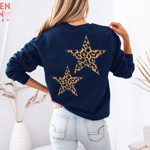 May include: Navy blue sweatshirt featuring two leopard print stars on the back. The sweatshirt has long sleeves and a crew neck. The VALEN TEEN design logo is visible in the upper left corner.