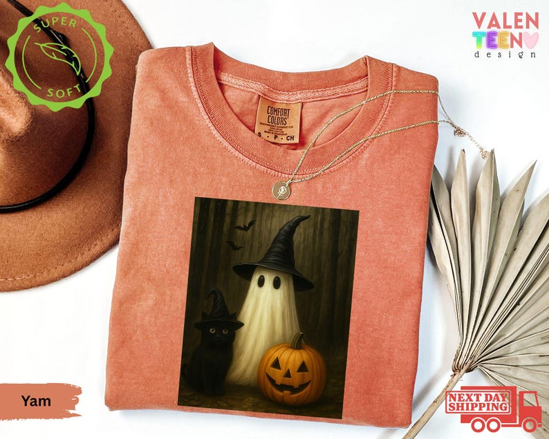 Puede incluir: Una camiseta de color coral con un gr&aacute;fico de Halloween. El gr&aacute;fico representa un fantasma con un sombrero de bruja, un gato negro y una calabaza tallada. La camiseta tiene cuello redondo y una etiqueta Comfort Colors.