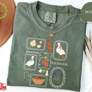 May include: Moss green t-shirt with a crew neck and short sleeves. The shirt features framed illustrations of a teapot, teacup, goose, strawberries, and lavender. A gold necklace rests on the shirt. The shirt has a tag that says "COMFORT COLORS".