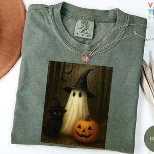 Puede incluir: Camiseta Comfort Colors verde musgo con un gr&aacute;fico de Halloween. El gr&aacute;fico incluye un fantasma con un sombrero de bruja, un gato negro y una calabaza tallada. Un delicado collar descansa sobre la camiseta. El texto "VALENTEEN design" es visible.