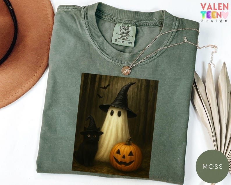 Puede incluir: Camiseta Comfort Colors verde musgo con un gr&aacute;fico de Halloween. El gr&aacute;fico incluye un fantasma con un sombrero de bruja, un gato negro y una calabaza tallada. Un delicado collar descansa sobre la camiseta. El texto "VALENTEEN design" es visible.