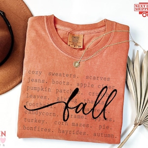 May include: A terracotta-colored crew neck t-shirt featuring black text listing fall-themed words like cozy sweaters, scarves, jeans, boots, apple cider, pumpkin patches, crunchy leaves, football, candy corn, caramel apples, turkey, and bonfires.