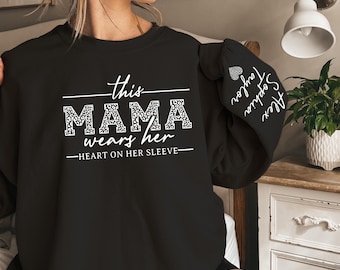 May include: Black sweatshirt with white text that reads "This Mama wears her heart on her sleeve." The text is surrounded by a leopard print design. The sleeves have white text that reads "Alex, Sophia, Taylor".