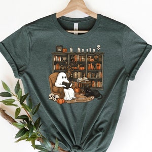 May include: A dark green t-shirt with a graphic of a ghost reading a book in a chair next to a black cat. The ghost is sitting in a chair in front of a bookshelf filled with books. There are pumpkins and skulls on the floor.
