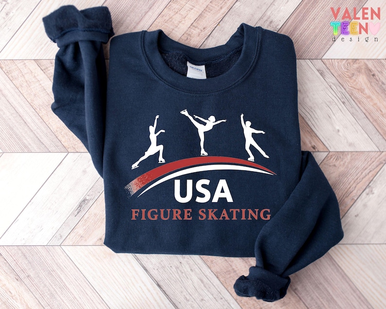 May include: Navy blue sweatshirt featuring three white figure skaters above a red and white curved design. The text "USA FIGURE SKATING" is printed below the design. The sweatshirt has a crew neck and long sleeves.