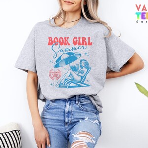 May include: A gray t-shirt with a graphic that reads "BOOK GIRL Summer" in red and blue. The graphic features a woman reading under a beach umbrella. The shirt is paired with blue jeans.