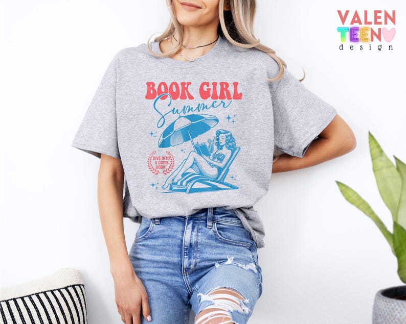 May include: A gray t-shirt with a graphic that reads "BOOK GIRL Summer" in red and blue. The graphic features a woman reading under a beach umbrella. The shirt is paired with blue jeans.