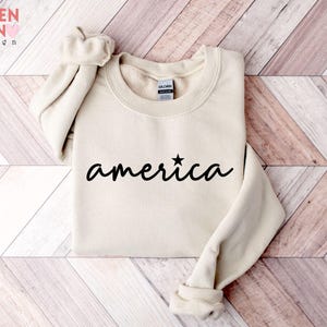 Puede incluir: Sudadera de color crema con la palabra "america" en escritura negra, una estrella reemplaza la letra "i". La sudadera tiene cuello redondo y mangas largas. La marca "VALENTEEN design" est&aacute; en la esquina superior izquierda.