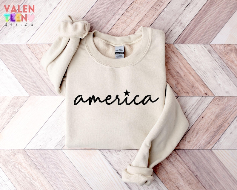 Puede incluir: Sudadera de color crema con la palabra "america" en escritura negra, una estrella reemplaza la letra "i". La sudadera tiene cuello redondo y mangas largas. La marca "VALENTEEN design" est&aacute; en la esquina superior izquierda.