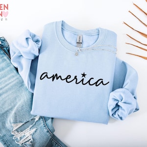 Puede incluir: Una sudadera azul claro con la palabra "america" en escritura negra, con una estrella encima de la "i". La sudadera se muestra con vaqueros rotos de lavado claro. La esquina superior izquierda tiene el texto "VALENTEEN design" en color arco&iacute;ris.