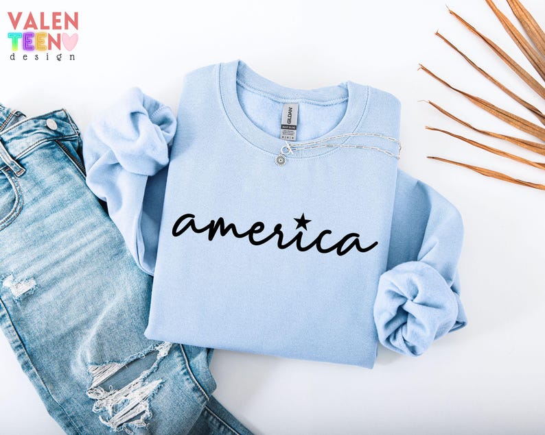 Puede incluir: Una sudadera azul claro con la palabra "america" en escritura negra, con una estrella encima de la "i". La sudadera se muestra con vaqueros rotos de lavado claro. La esquina superior izquierda tiene el texto "VALENTEEN design" en color arco&iacute;ris.