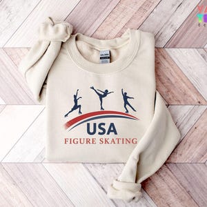May include: A cream-colored sweatshirt featuring a graphic of three figure skaters in silhouette, with the text "USA FIGURE SKATING" in navy and red. The sweatshirt has a crew neck and long sleeves.