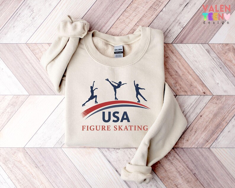 May include: A cream-colored sweatshirt featuring a graphic of three figure skaters in silhouette, with the text "USA FIGURE SKATING" in navy and red. The sweatshirt has a crew neck and long sleeves.
