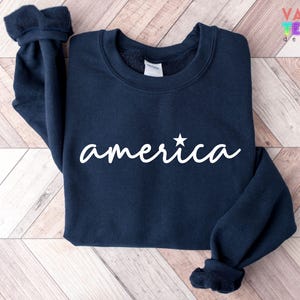 Puede incluir: Sudadera azul marino con la palabra "america" en escritura blanca, con una estrella sobre la "i". La sudadera tiene cuello redondo y mangas largas con pu&ntilde;os acanalados. La marca "VALENTEEN design" est&aacute; impresa en la esquina superior derecha.