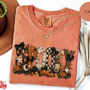 May include: A burnt orange short-sleeve t-shirt featuring the word SPOOKY in large, patterned letters. The letters include designs like plaid, leopard print, and ghost motifs, accented with small black bows, pumpkins, stars, and moons.