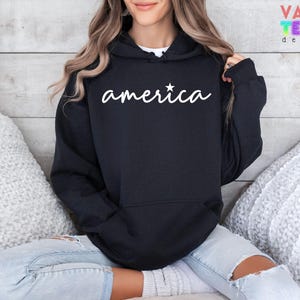 Puede incluir: Sudadera con capucha negra con la palabra "america" en escritura blanca y una estrella blanca. La sudadera tiene un bolsillo delantero y una capucha con cord&oacute;n. El dise&ntilde;o est&aacute; en la parte delantera de la sudadera.