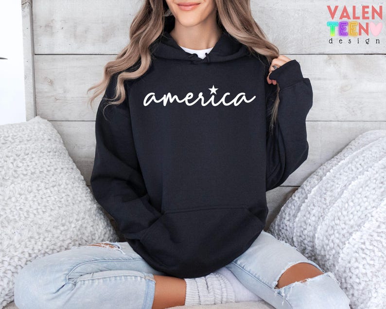 Puede incluir: Sudadera con capucha negra con la palabra "america" en escritura blanca y una estrella blanca. La sudadera tiene un bolsillo delantero y una capucha con cord&oacute;n. El dise&ntilde;o est&aacute; en la parte delantera de la sudadera.