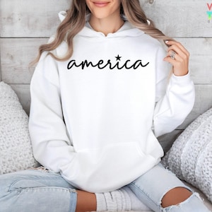Puede incluir: Sudadera blanca con la palabra "america" en escritura negra, con una estrella encima de la "i". La sudadera tiene un bolsillo delantero y una capucha. La persona lleva vaqueros azul claro.