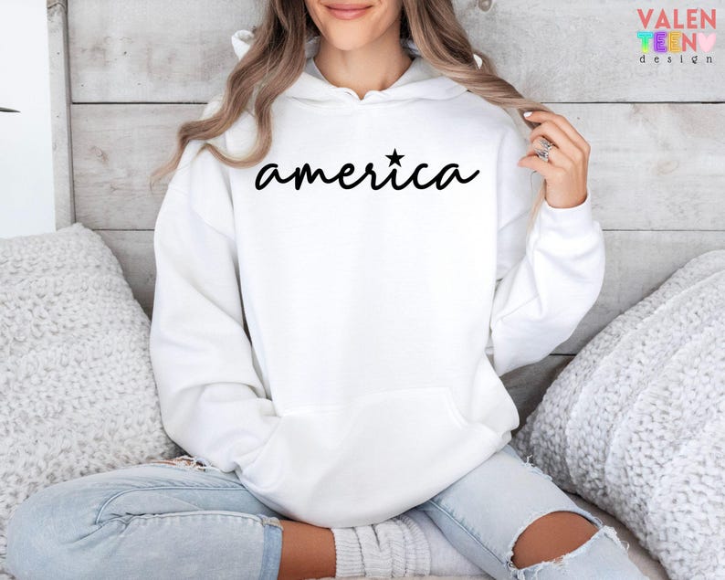 Puede incluir: Sudadera blanca con la palabra "america" en escritura negra, con una estrella encima de la "i". La sudadera tiene un bolsillo delantero y una capucha. La persona lleva vaqueros azul claro.
