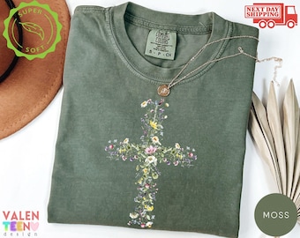 May include: A moss green short-sleeve t-shirt featuring a cross graphic composed of various colorful wildflowers. The shirt has a crew neckline and a Comfort Colors brand label on the inside collar. A thin gold-toned necklace rests on the fabric.