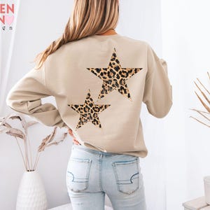 May include: Beige sweatshirt with two leopard print stars on the back. The sweatshirt has long sleeves and a crew neck. The text "VALEN TEEN design" is visible in the upper left corner. The stars are brown, black, and tan.