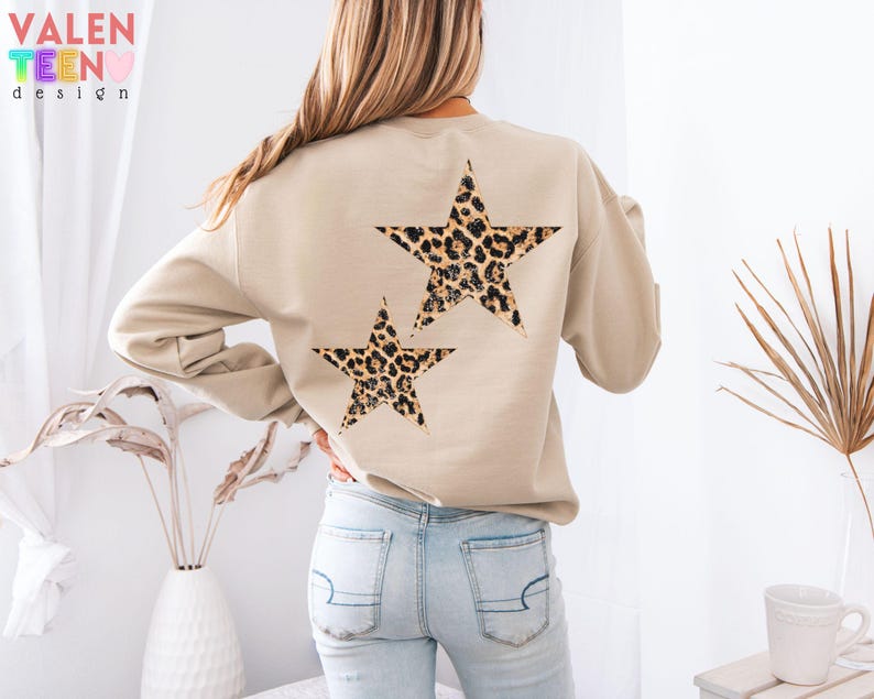 May include: Beige sweatshirt with two leopard print stars on the back. The sweatshirt has long sleeves and a crew neck. The text "VALEN TEEN design" is visible in the upper left corner. The stars are brown, black, and tan.