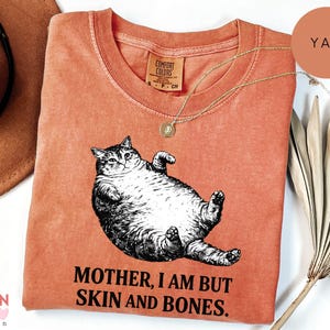 May include: A coral Comfort Colors sweatshirt with a black and white cat illustration and the text "MOTHER, I AM BUT SKIN AND BONES". A gold necklace is draped across the shirt. The shirt is a warm, earthy tone, with a relaxed fit.