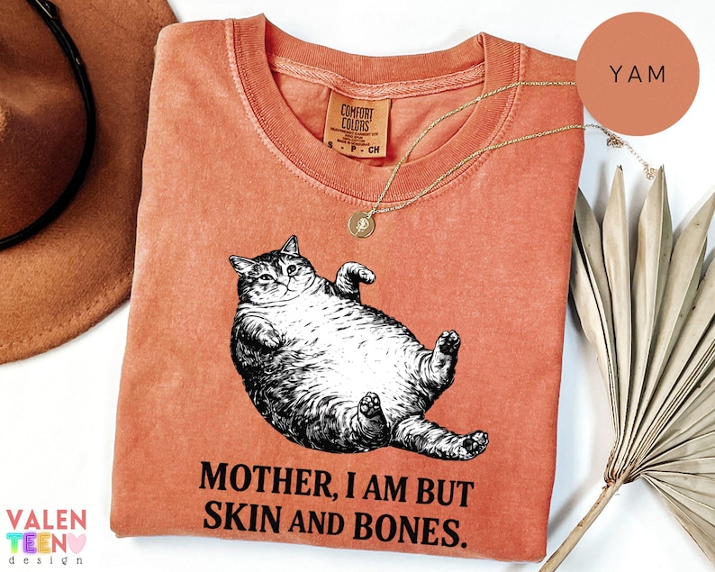 May include: A coral Comfort Colors sweatshirt with a black and white cat illustration and the text "MOTHER, I AM BUT SKIN AND BONES". A gold necklace is draped across the shirt. The shirt is a warm, earthy tone, with a relaxed fit.