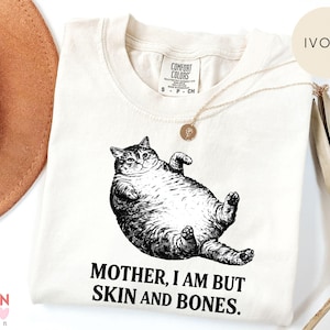 May include: Cream-coloured t-shirt with a black and white drawing of a cat on its back. The text "MOTHER, I AM BUT SKIN AND BONES" is printed beneath the cat. The shirt has a round neck and short sleeves.