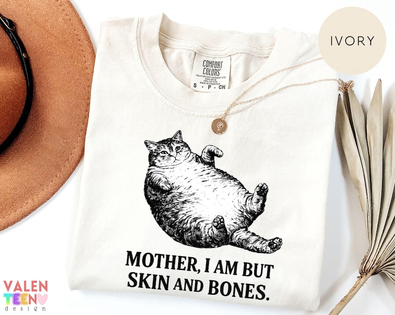 May include: Cream-coloured t-shirt with a black and white drawing of a cat on its back. The text "MOTHER, I AM BUT SKIN AND BONES" is printed beneath the cat. The shirt has a round neck and short sleeves.